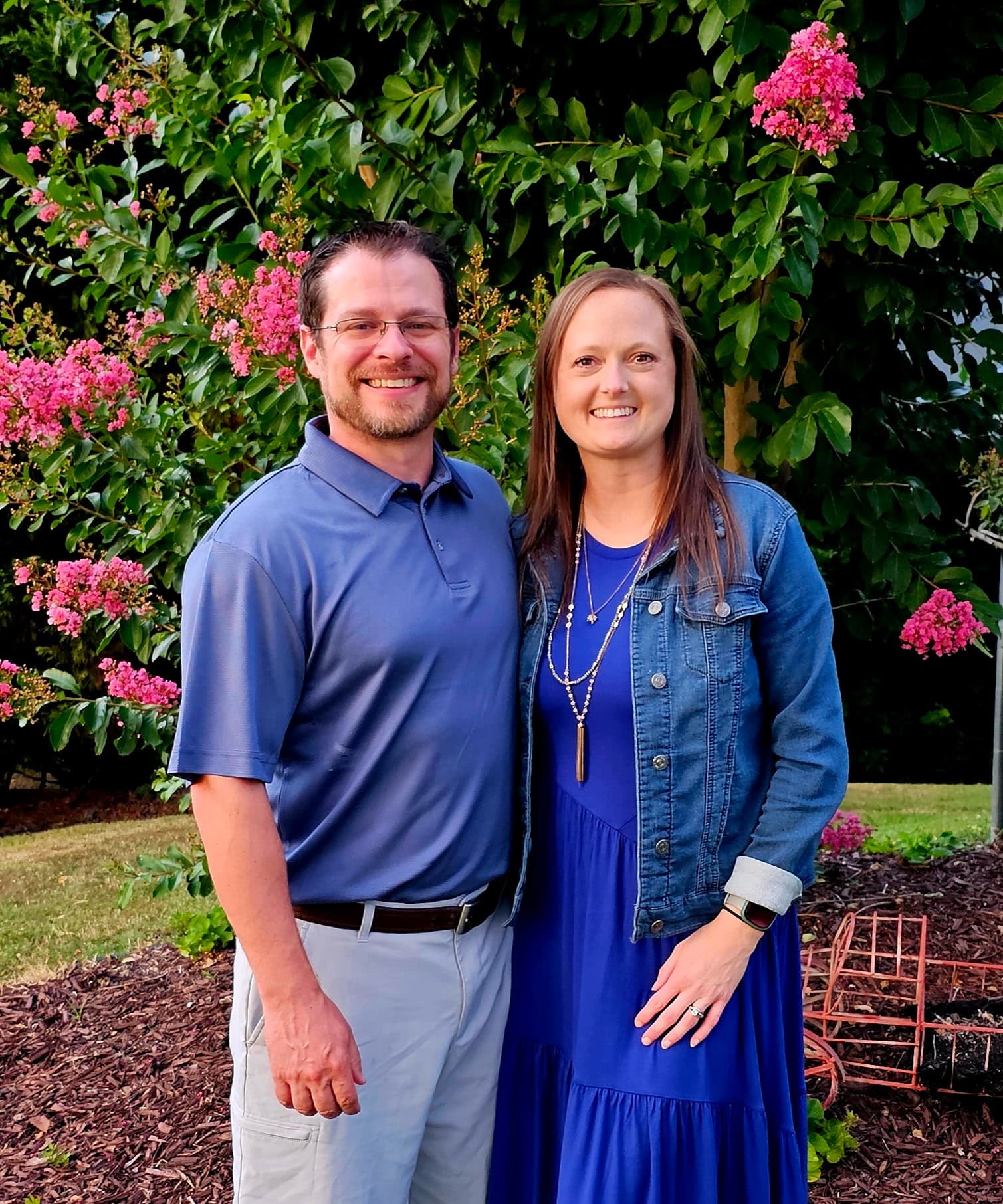 Smiling couple in blue outfits posing in front of a lush pink flowering bush.