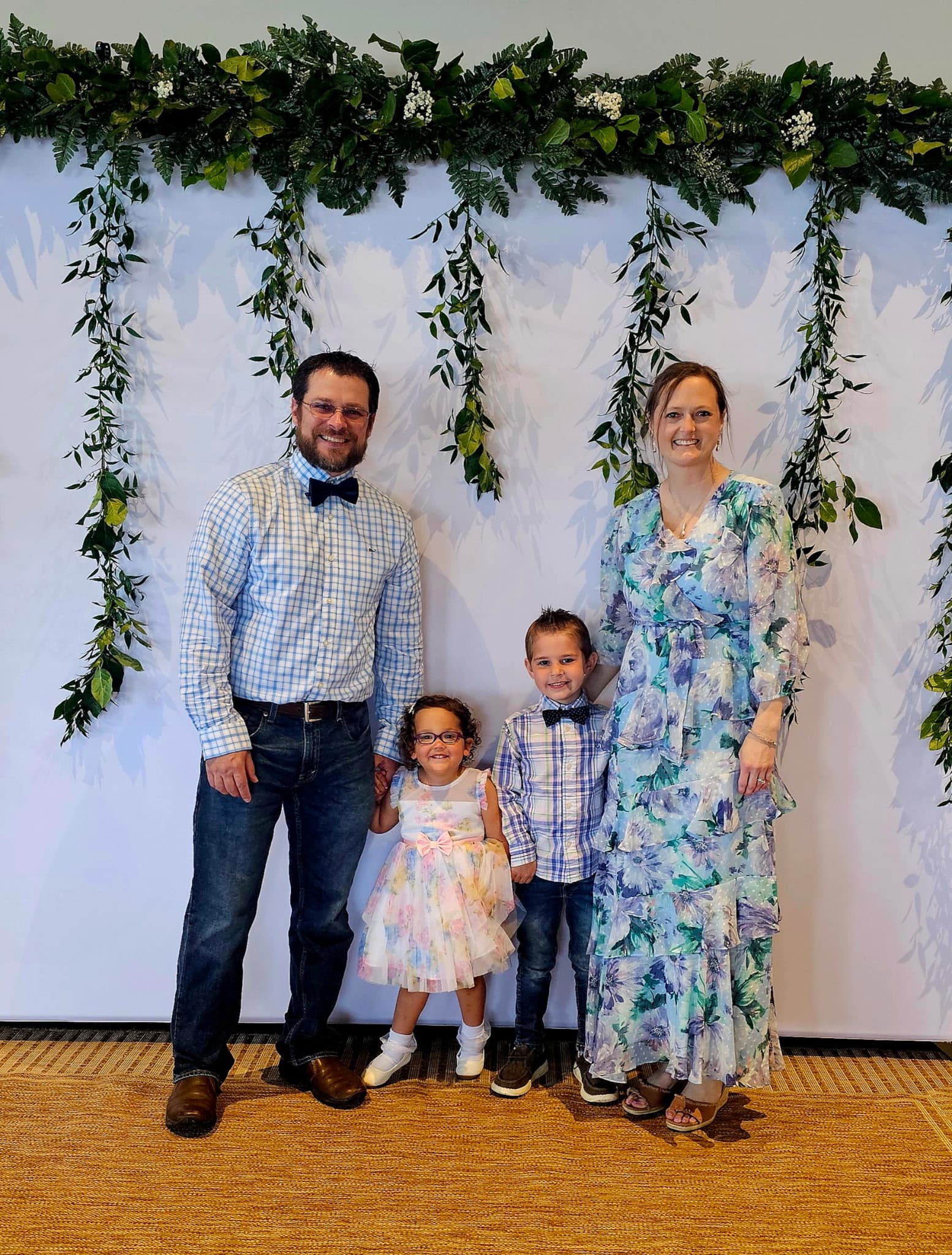 Family of four smiling in front of a white backdrop with hanging green leafy garlands.