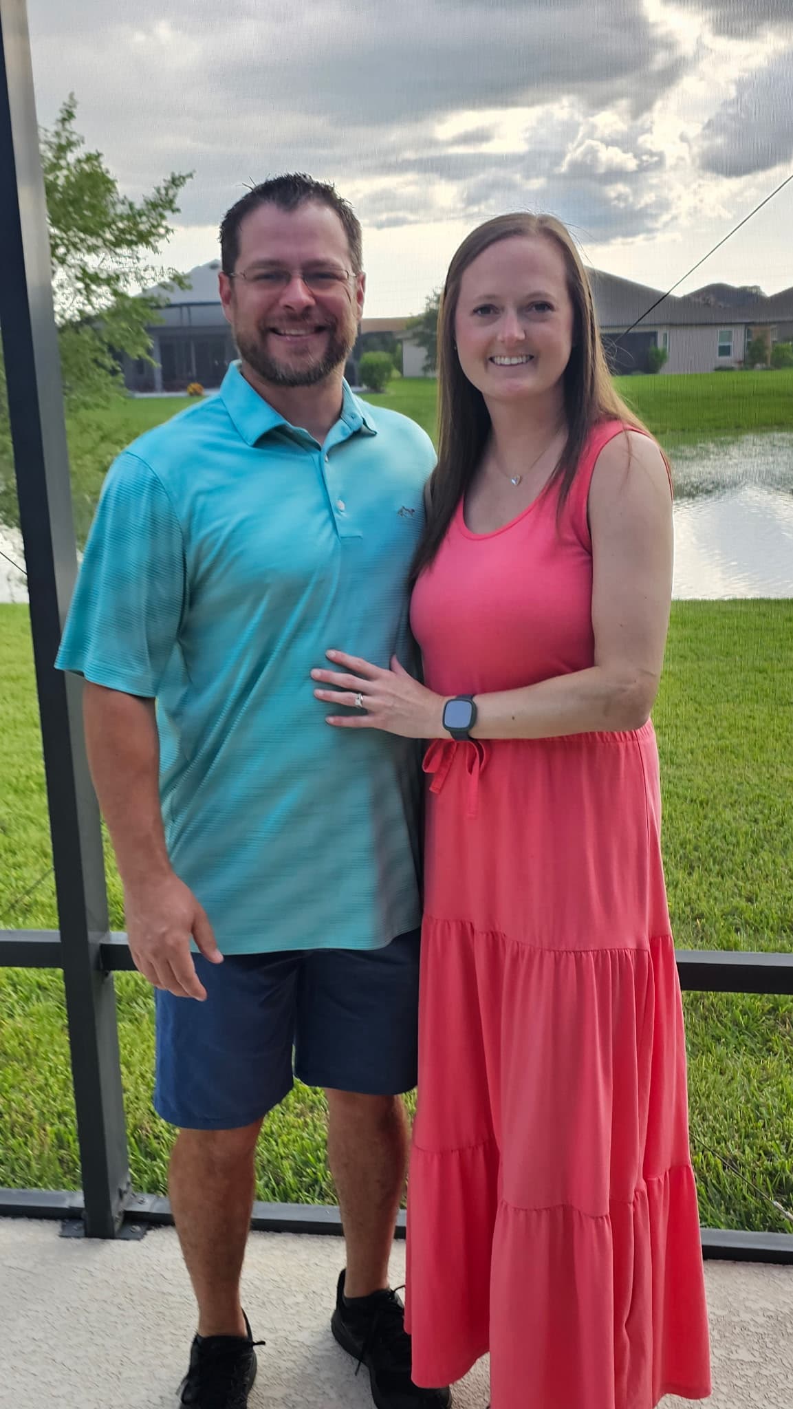 Smiling man in blue polo and woman in pink dress pose together on a patio.