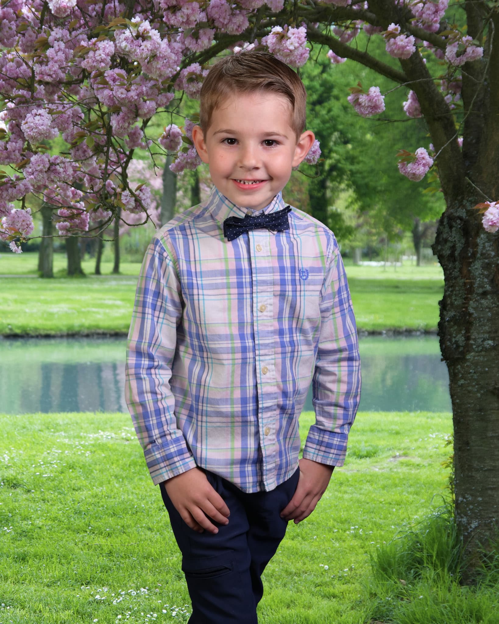 Smiling young boy in a plaid shirt and bow tie under pink cherry blossoms.