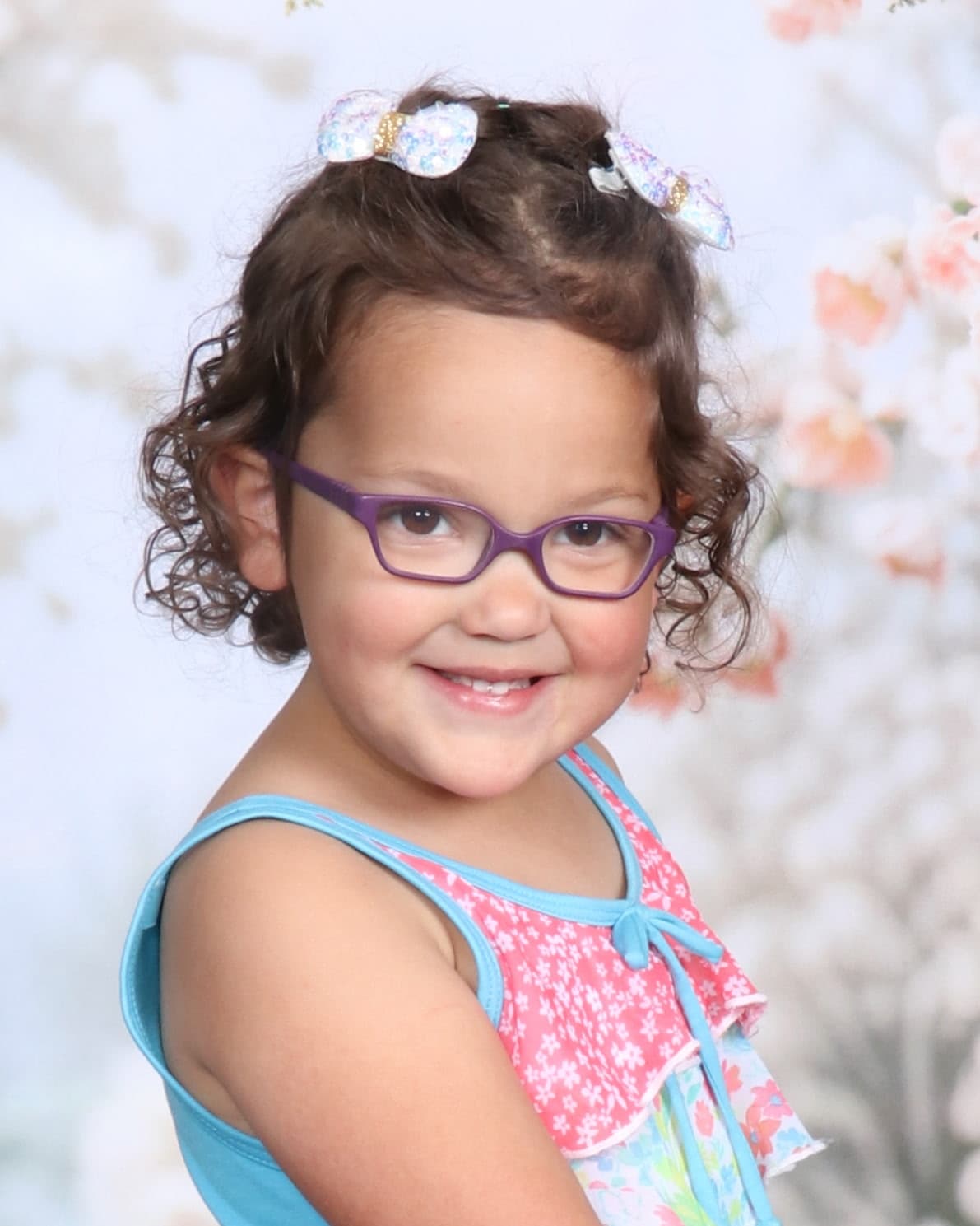 Smiling young girl with curly hair, purple glasses, and sparkly bows against a floral backdrop.