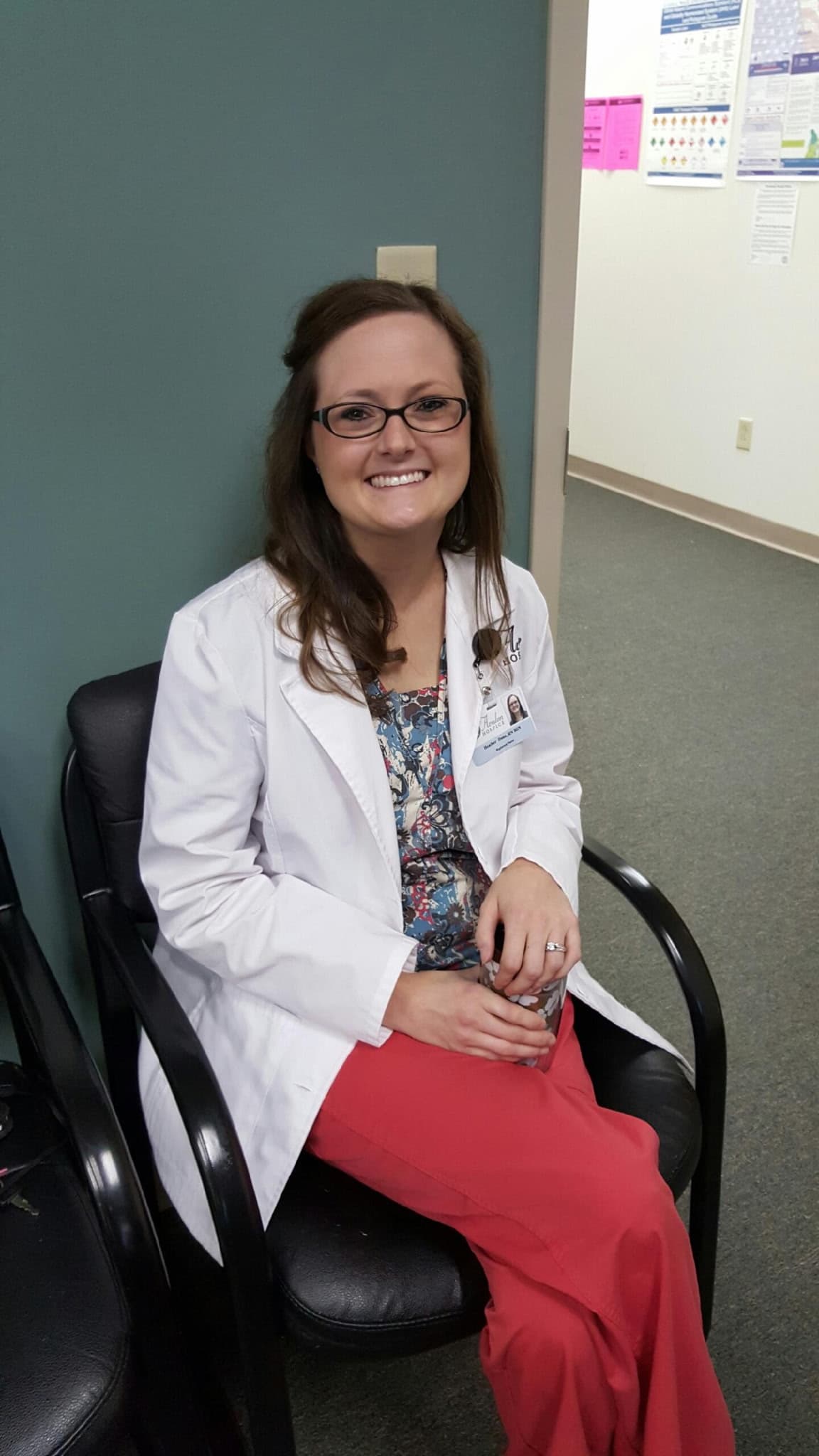 Smiling woman in glasses, white lab coat, and red scrubs sitting in a black chair.
