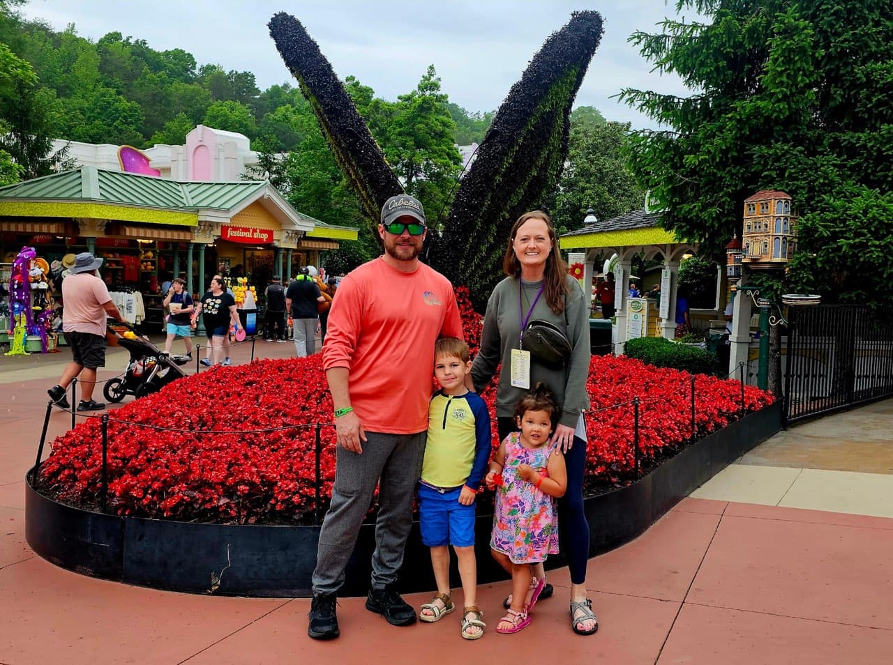 Family of four posing in front of a red flower bed and large butterfly topiary.