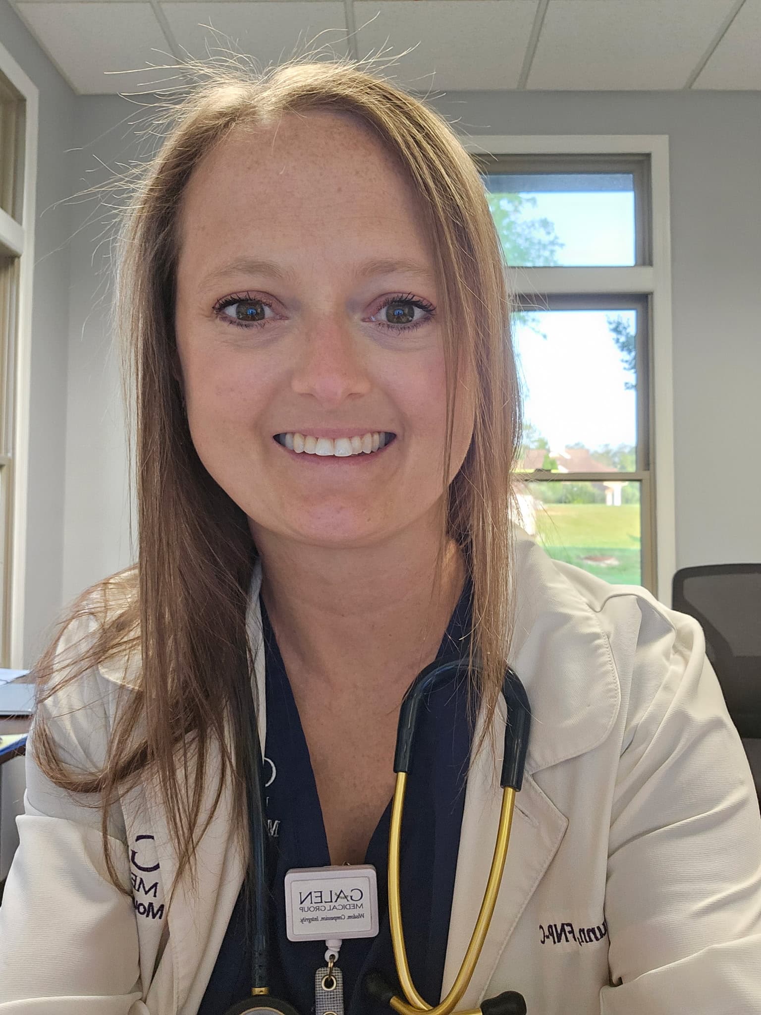 Smiling female medical professional wearing a white lab coat and stethoscope in a bright office.